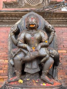 A large dark stone multi-armed Narasimha (lion-headed deity) sculpture adorned with marigold flower offerings and vermillion, set against a carved temple wall in Patan Durbar Square, Nepal. 