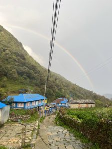 Village scene with houses featuring blue roofs nestled against a lush, green hillside. A rainbow arches across the sky, creating a serene, picturesque atmosphere.