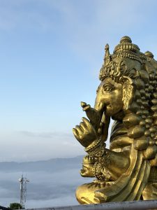 Golden statue of Ganesha against a clear blue sky and misty mountains. The intricate details and serene expression convey tranquility and spirituality.