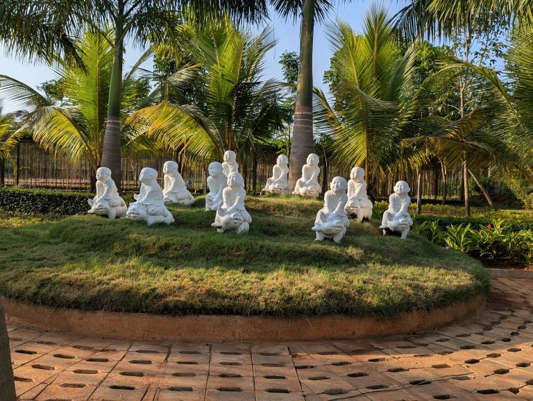 White stone monk figurines seated on a grass mound surrounded by tall palm trees in a garden setting, with warm sunlight filtering through the leaves.