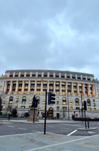 A wide shot of a massive, classical stone building with columns and many glowing windows at dusk. A paved city square, a traffic light, and a statue are visible in the foreground under a cloudy sky. 