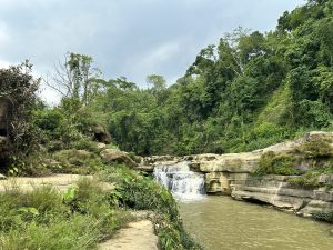 Nafakhum waterfall flows over layered rocks surrounded by lush green foliage under a cloudy sky, conveying a peaceful and natural landscape.
