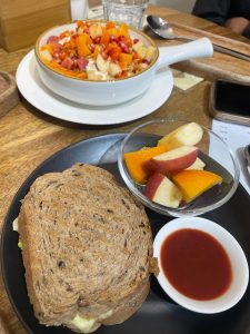 Whole-grain sandwich, fresh fruit slices, and red sauce on a black plate, with a bowl of yogurt topped with papaya and pomegranate. 