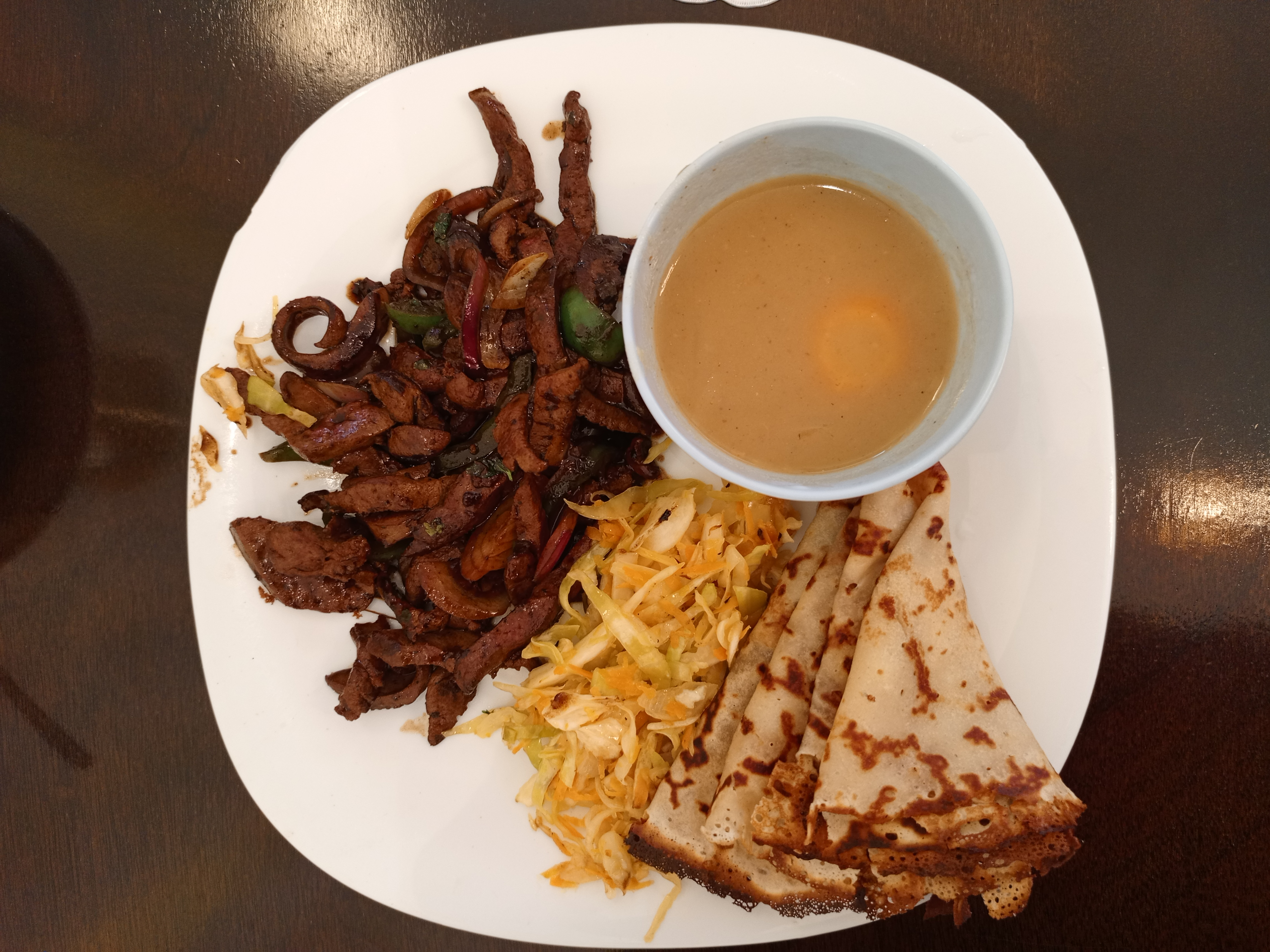 A white plate contains a serving of thin, sautéed strips of liver mixed with sliced onions and green bell peppers. It is served alongside a portion of shredded cabbage and carrot salad, several folded crepes, and a light brown dipping sauce in a small white bowl.