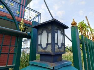 An ornate dark blue metal gate lantern on a decorative teal fence post with a colourful Nepali house facade behind it. 