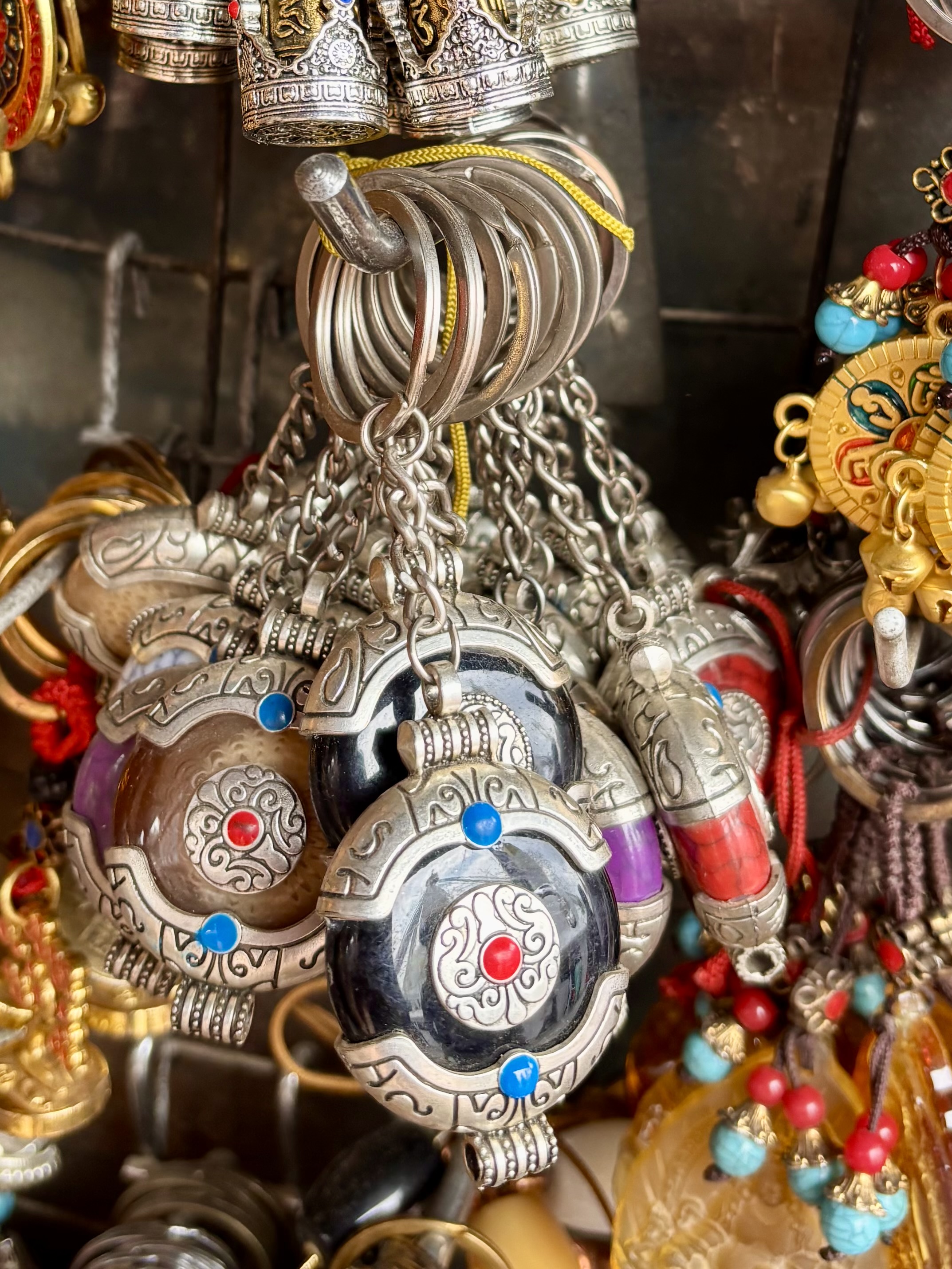 Ornate keychains with intricate silver designs and colorful stones hang from a metal rod in a vibrant market setting.