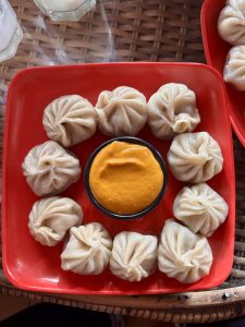 A dozen plump steamed momos arranged in a circle around a small bowl of golden-orange achar (chili dipping sauce) on a red square plate.