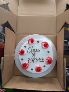 White celebration cake in a box with “Class of 2023–26” written on top, decorated with red cream swirls, cherries, and chocolate dots, with drinks and utensils nearby.

