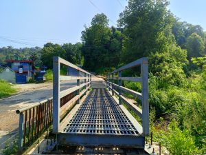 A rustic steel-grated footbridge leads toward a tree-lined road in a quiet Nepali hillside town under clear morning skies. 