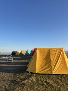 A row of colorful tents, including yellow, blue, and red, set up on a grassy hill under a clear blue sky, creating a peaceful, early morning camping scene.