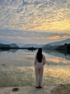A scenic shot from behind of a person looking out over a calm, reflective lake during a vibrant sunset, with golden and orange clouds filling the sky and rolling hills in the background. 
