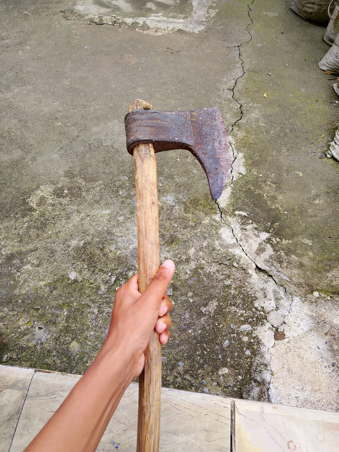 A hand grips the wooden handle of a worn, rusted axe above a cracked concrete floor.