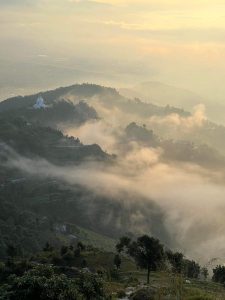High-angle landscape of a white dome structure on a green hill ridge, partially covered by thick, swirling morning fog and mist lit with warm golden sunlight. 