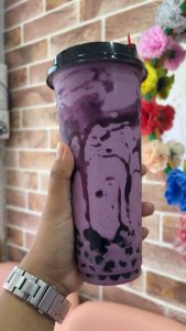 A hand holding a tall purple bubble tea with black tapioca pearls against a brick wall backdrop. Bright paper flowers create a cheerful ambiance.