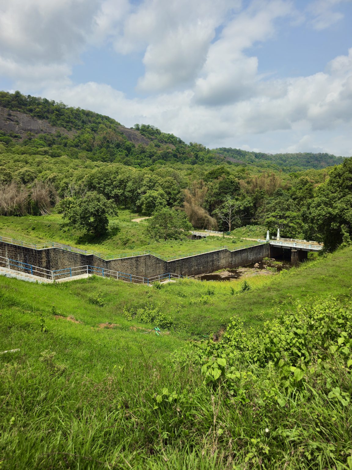 A panoramic view of green hills, forest vegetation, and the lower section of the dam structure. The rich shades of green and dramatic cloud-filled sky showcase the natural beauty of the Western Ghats. Captured at Parambikulam Tiger Reserve, Palakkad District, Kerala.