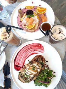 Top-down view of a brunch table featuring a face-shaped pancake with fruit, a savory toast with microgreens and a red sauce swirl, and two iced coffees with curly black straws.