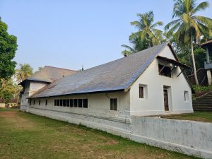 A long traditional Kerala temple building with a sloping tiled roof stands amid coconut trees and open grounds. The simple architecture reflects the temple's heritage and peaceful surroundings. Captured at Niramkaitha Kotta Bhagavathi Temple, Vallikunnu North, Malappuram.