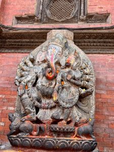 A multi-armed stone Ganesh sculpture with consort, adorned with marigold flowers and vermillion powder offerings, set into a temple wall in Patan, Nepal.