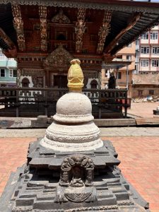 A whitewashed Buddhist chaitya with a golden finial on a stepped black stone plinth with a niche housing a small deity figure, in front of a decorated Newari pagoda temple, Nepal. 
