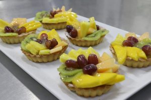 Assorted fruit tarts topped with kiwi, grapes, mango, pineapple, and melon on a white rectangular plate. 
