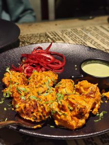 Juicy tandoori chicken tikka garnished with fresh coriander, served with pickled red onions and mint chutney on a dark platter 