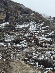 A vertical, wide shot of a lone hiker seen from behind, walking away along a narrow, rocky dirt path. The trail ascends into a steep, rugged mountain slope covered with large boulders, dark soil, patches of dry brown vegetation, and scattered remnants of white snow.
