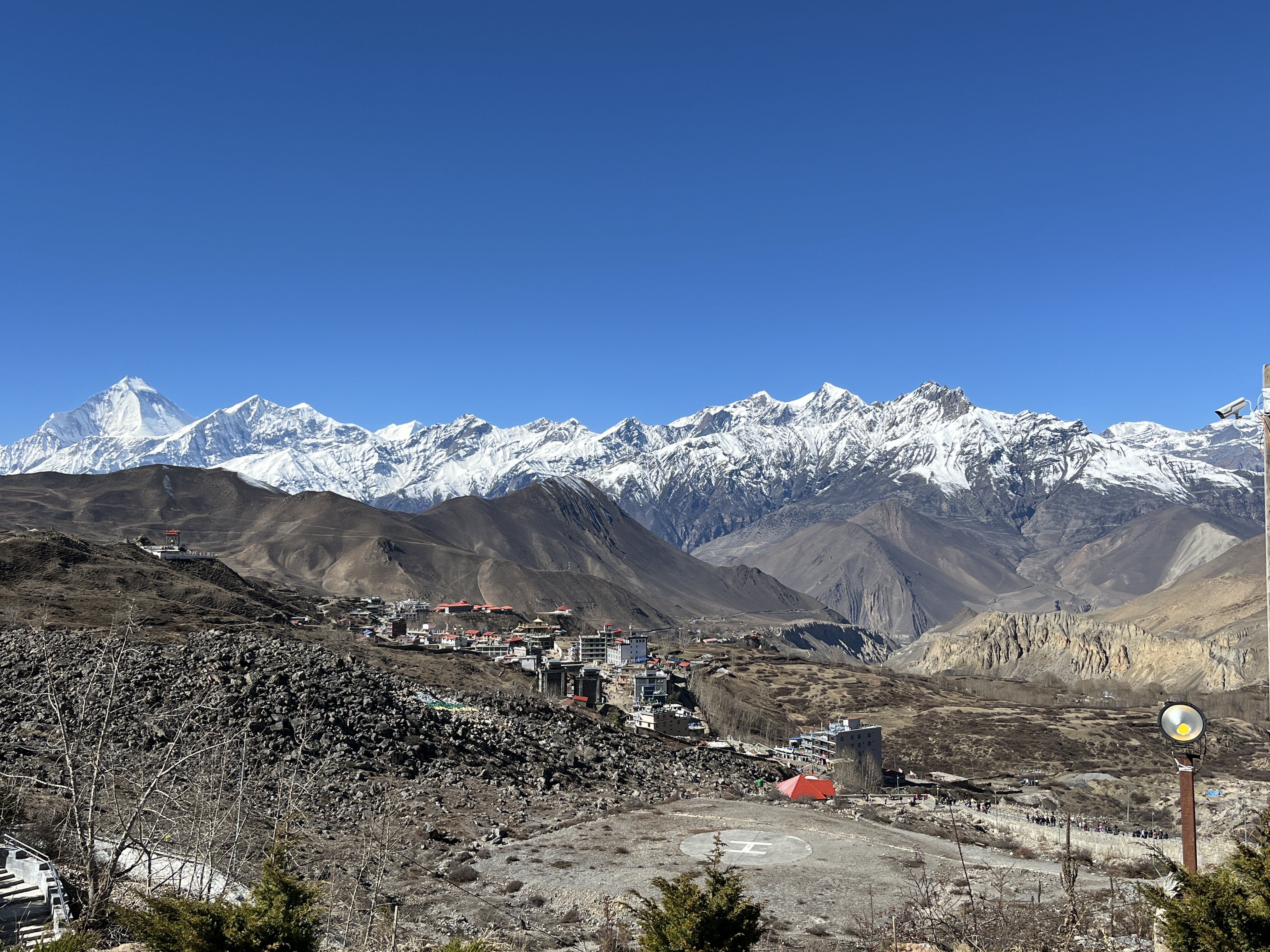 This landscape photo from Mustang, Nepal shows a high-altitude village in a dry, barren area with the snow-covered Himalayas in the background. The colorful houses reflect how people live in and adapt to this harsh, rugged environment.