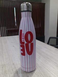 Stainless steel bottle with pink and white stripes and bold red letters spelling “LOVE.” It stands on a light wooden table in a modern room.
