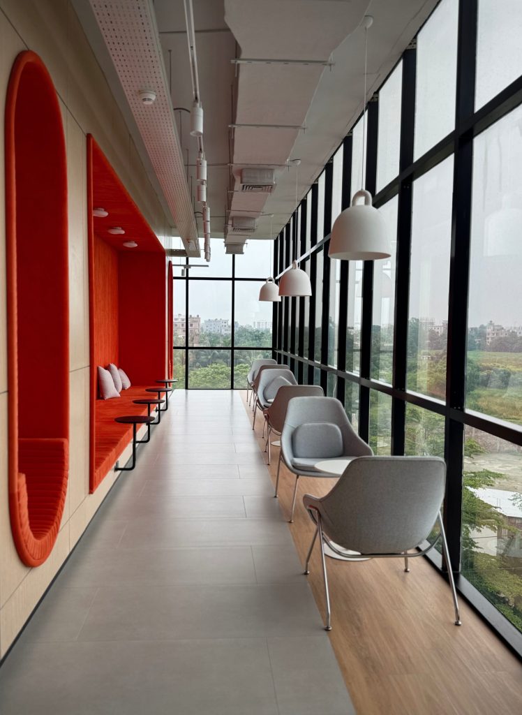 Modern lounge with vibrant orange seating alcoves on one side and sleek grey chairs with white tables on the other, all by tall glass windows.