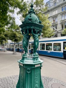 A beautiful dark green Wallace Fountain made of cast iron stands in a paved square in Zurich, Canton of Zurich, Switzerland. Four elegant female statues support its decorative dome, while a modern blue-and-white tram passes through the blurred background, blending the city's historic charm with modern urban life.