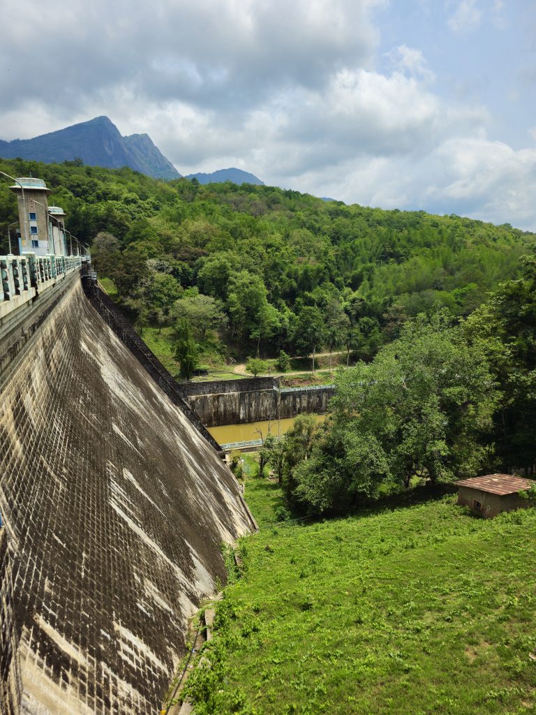 A scenic photograph of a dam surrounded by lush green forests and distant mountain peaks. The strong lines of the dam contrast beautifully with the natural landscape, highlighting both engineering and nature. Captured at Parambikulam Tiger Reserve, Palakkad District, Kerala.