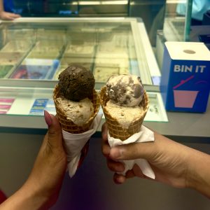 Two hands holding double-scoop waffle ice cream cones — one with chocolate and caramel, one with cookies and cream — at an ice cream parlor.
