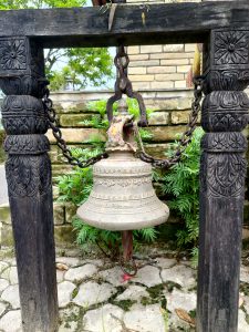 An aged brass temple bell suspended by iron chains from a carved black wooden frame at a Nepali shrine. 
