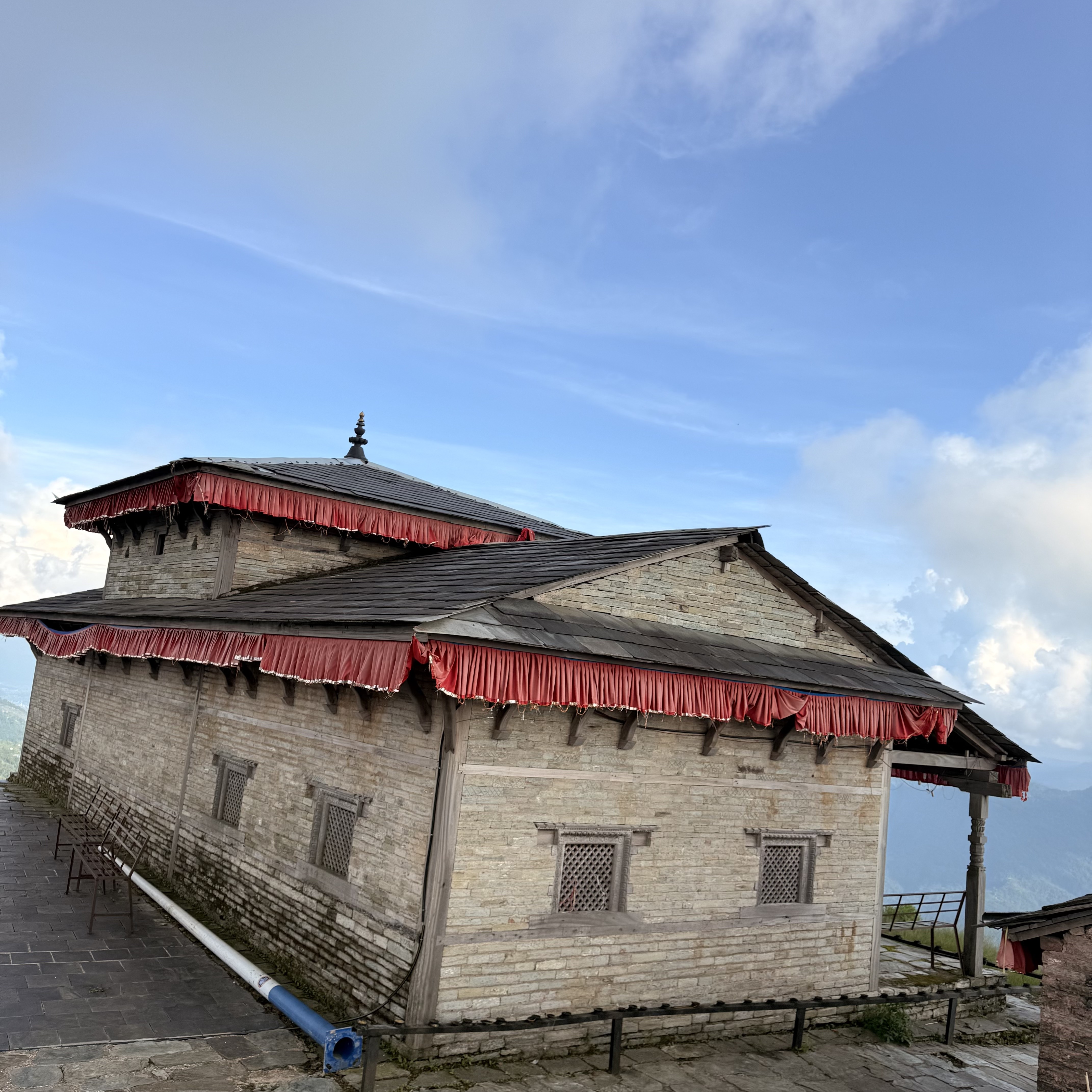 Traditional stone building with a dark tiered roof and red trim, featuring carved lattice windows under a blue sky with white clouds.