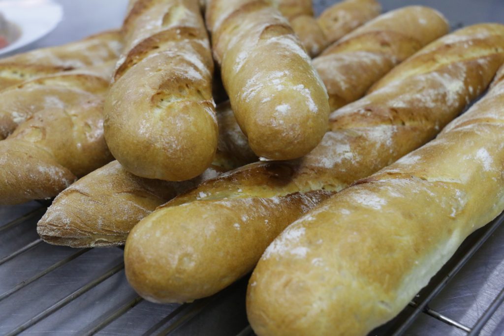Many freshly baked golden-brown baguettes bread loaves dusted with flour rest on a metal cooling rack. It shows crisp crusts with diagonal scoring along the tops.