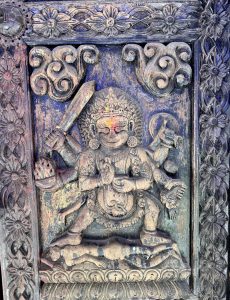 Intricate wood carving on a temple door panel depicting a multi-armed Hindu deity (possibly Mahakali or Bhairava) holding a sword, painted in blue with fresh vermillion offerings. 