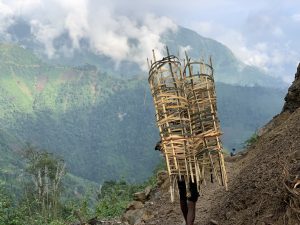 A person walks along a dirt mountain path carrying several large cylindrical bamboo structures on their back, with green hills and cloud‑covered mountains in the background. 