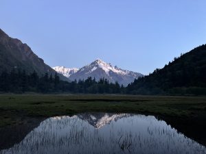 Snow-capped mountain under a clear blue sky, reflected in a still pond. Dense forest lines the base, conveying tranquility and natural beauty.