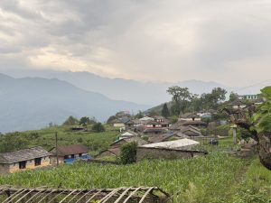 A misty panoramic view of a traditional Nepali hillside village with stone-roofed houses, maize fields, and layered mountain ranges under a cloudy sky, Nepal. 
