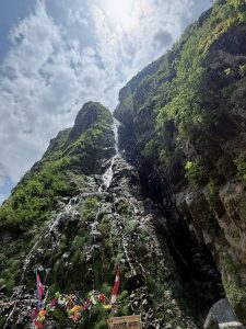 Mountain Octopus Waterfall cascades down a rocky cliff, surrounded by lush greenery. Bright sky overhead. 