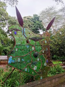 A large public monument crafted from green and brown mosaic glass panels configured into an abstract geographic silhouette of Kenya, accented with stylized glass faces and two spear-like elements at the top. 
