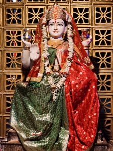 A full view of a richly decorated idol of the goddess Annapurna, seated against an ornate, patterned backdrop. The vibrant attire, floral garlands, and detailed craftsmanship reflect the artistic and spiritual traditions of Kolkata. Captured in Kolkata, West Bengal.