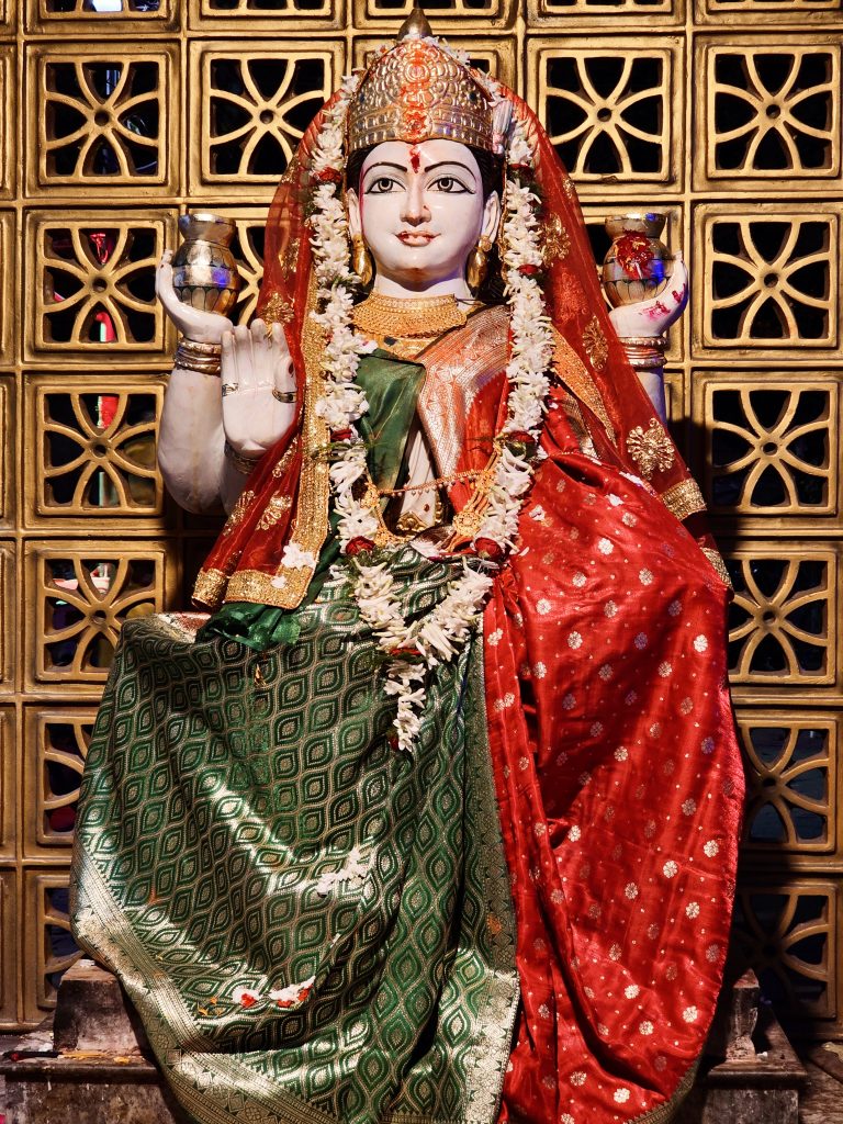 A full view of a richly decorated idol of the goddess Annapurna, seated against an ornate, patterned backdrop. The vibrant attire, floral garlands, and detailed craftsmanship reflect the artistic and spiritual traditions of Kolkata. Captured in Kolkata, West Bengal.