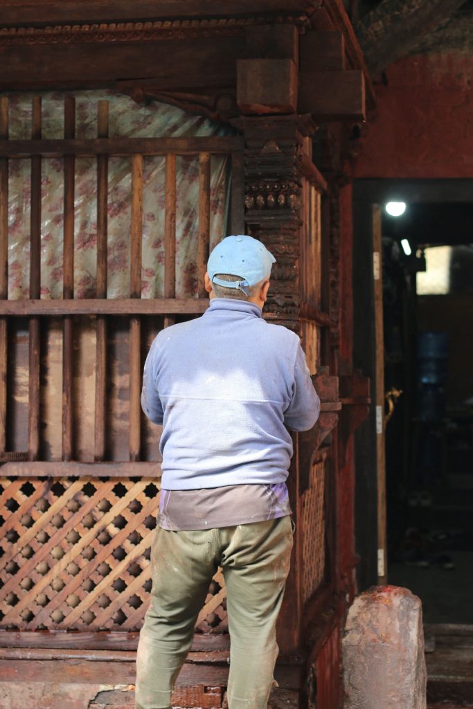 Man in blue cap and jacket working on intricate wood carvings on a traditional structure.