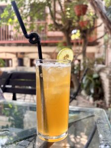 A tall iced passionfruit drink with a lime wheel garnish and a black spiral straw on a glass-top table in a leafy outdoor café courtyard. 