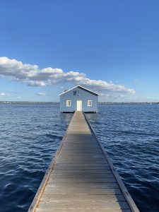 The “Blue House” is a vibrant, iconic wooden structure resting on a narrow timber jetty over the calm blue waters of the Swan River in Crawley, Perth.
