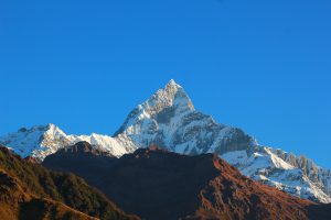 A majestic snow capped Machapuchare mountain peak stands against a clear blue sky. The foreground shows dark, rugged hills, creating a striking contrast. 