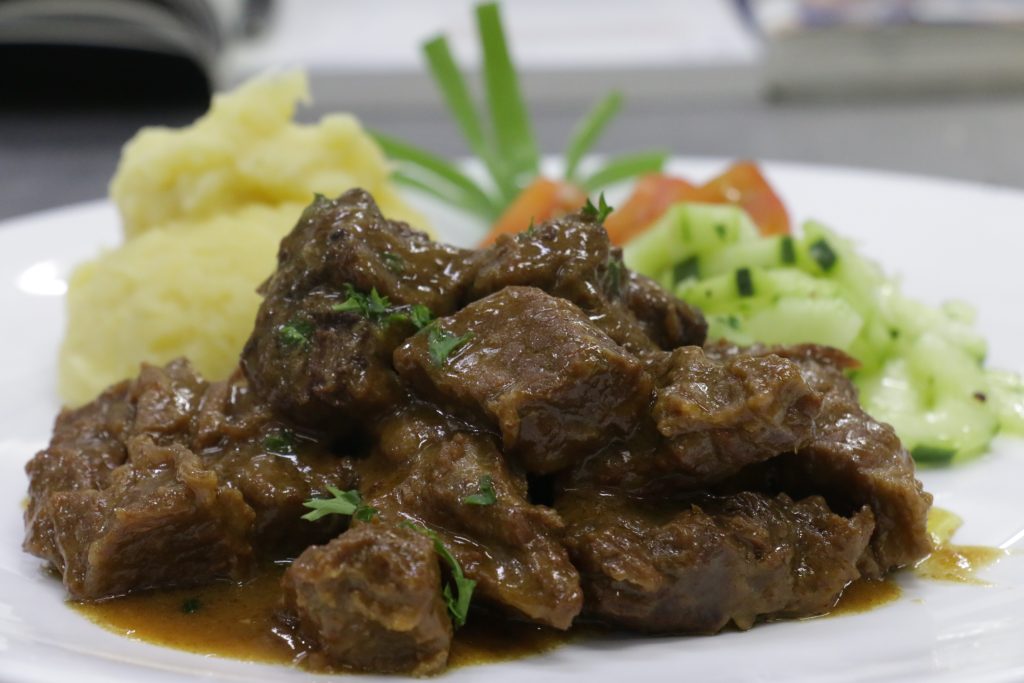 Chunks of meat in brown gravy served on a white plate with mashed potatoes, diced cucumber salad, tomato wedges and green onion garnish in the background.