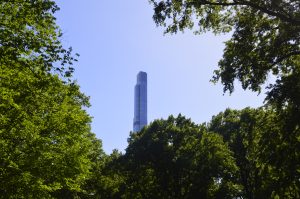 A tall glass skyscraper rising above green trees under clear blue sky