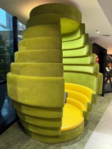 A bright, round, green and yellow pod-like chair with layered, cushioned walls, offering a cozy, futuristic retreat in a modern indoor setting.